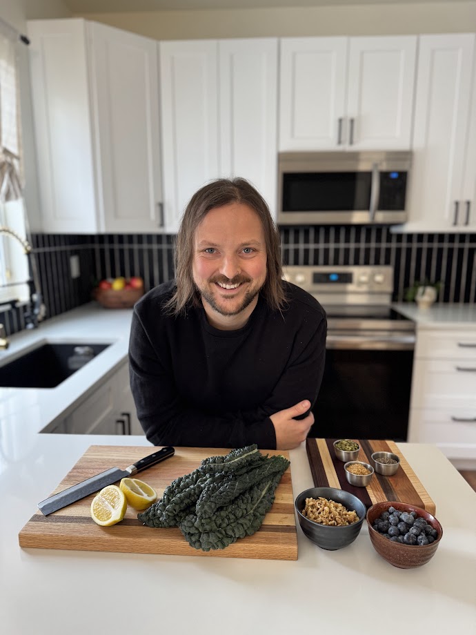 A smiling individual with shoulder-length hair stands in a modern kitchen, leaning on a white counter. In front of them are fresh kale leaves, a halved lemon, a knife on a wooden cutting board, and small bowls containing blueberries, walnuts, pumpkin seeds, and chia seeds. The background features white cabinets and stainless steel appliances.